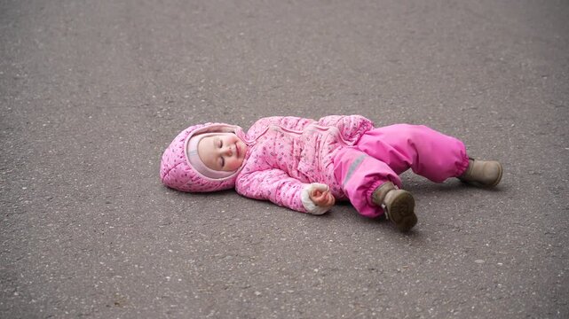 Toddler in pink winter outerwear resting or having tantrum lying flat on drab asphalt surface outdoors