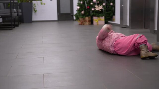 Little child in warm winter clothing playfully tumbles and rolls on floor of decorated lobby with Christmas trees and presents.