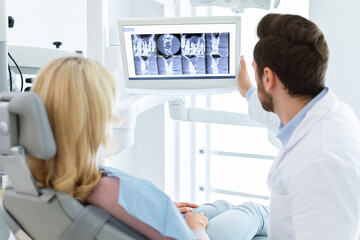 Obraz premium Modern dental clinic concept. Brunette man dentist having conversation with female patient sitting in dental chair, poitning at LED display, showing results of treatment or explaining procedure