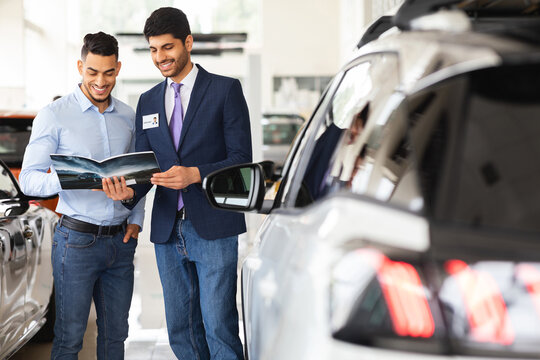 Middle-eastern guy having conversation with sales assistant in auto showroom. Handsome car dealer showing catalogue with various luxury autos, selling nice automobiles in brand new salon