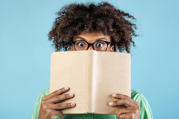 Portrait of emotional african american guy in glasses covering face with book, hiding behind it with eyes wide open, standing over blue studio background, closeup