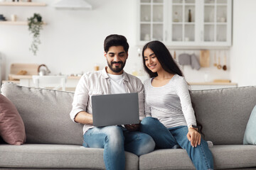Fototapeta premium Covid-19 and self-isolation at home, online conference, great website, offer or ad. Happy millennial muslim male and female looking at laptop, chatting with family in living room interior, copy space