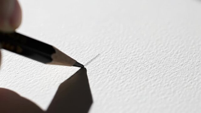 Close up shot of a person's hand holding a black pencil and drawing a straight line on a white textured paper creating a shadow effect under bright daylight