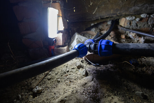 Water pipes connecting with blue compression fittings on a dirt floor in a dark basement or crawl space, illuminated by a work light, highlighting household plumbing maintenance.