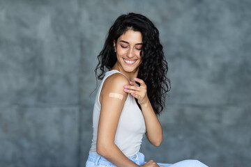 Fototapeta premium Coronavirus vaccination concept. Young Caucasian brunette woman showing adhesive bandage on her arm, getting vaccinated against covid-19 on grey background. Immunization for viral disease