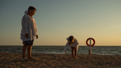 Brother and sister draw in wet sand beside calm sea at golden hour, lifebuoy in background as warm...
