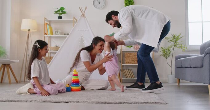 First steps of baby with father helping, barefoot toddler walking to happy mother at home living room with teepee and toys. Smiling parents kiss toddler, celebrate milestone of child with joy