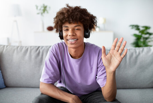 Black teenager talking to his friend or family online, wearing headphones and waving at camera from home. Cheerful African American adolescent having web conference, chatting to someone remotely