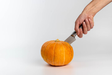 Hand holding a knife is positioned above a bright orange pumpkin on a white background, preparing for cutting or carving during autumn or Halloween festivities
