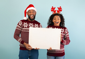Christmas ad. Happy black spouses holding empty poster, standing over blue background. Woman posing in deer horns and man in Santa hat, smiling to camera, banner