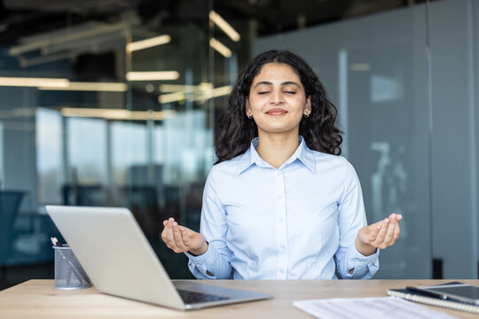 Young businesswoman finding peace and calm with eyes closed, hands in gyan mudra, practicing meditation and mindfulness right at her office desk to manage stress