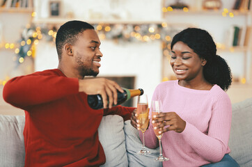 Obraz premium A man pours sparkling drink into glasses held by a woman as they sit on a couch. The room is decorated for a celebration with soft lights. They are enjoying a fun moment together.
