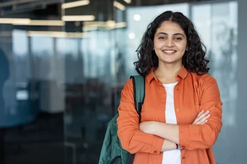 Fototapete Altes Krankenhaus Beelitz Young indian woman with a backpack smiling confidently, posing with crossed arms in a contemporary office or university setting, representing education, professionalism, and success  © Liubomir
