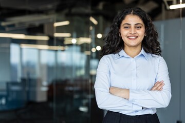 Young professional businesswoman standing with a confident smile and crossed arms in a contemporary corporate office building, symbolizing success and professional growth
