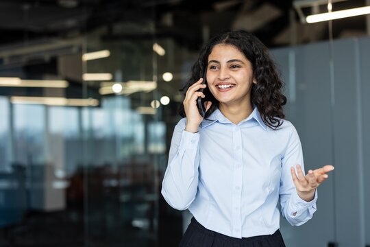 Indian businesswoman standing in a modern office, enjoying a pleasant conversation on her mobile phone, actively communicating while expressing happiness and professionalism