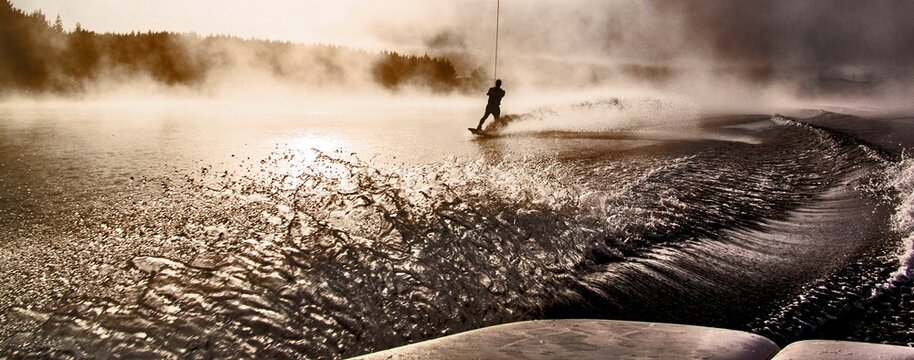 A person is wakeboarding on a lake during sunset. Mist rises from the water. Ripples form as the skier moves. The scene has trees in the background creating a natural setting.