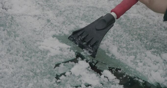 A man cleans snow and ice from his car windshield with a scraper on a cold winter morning. A man prepares a car for safe driving during heavy snowfall and improves visibility for vehicles.