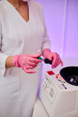 Woman in white lab coat and pink gloves holding a test tube near a centrifuge under soft purple lighting in a clinical setting