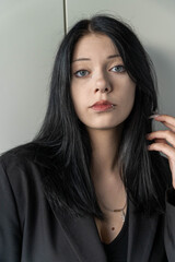 Naklejka premium Young woman with long black hair wearing a black blazer poses thoughtfully against a light background, showcasing her piercing and subtle makeup in a portrait setting