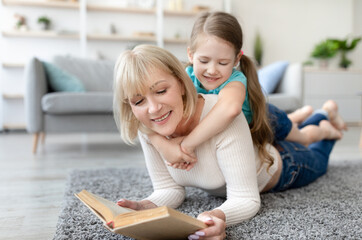 Naklejka premium Loving grandmother teaching granddaughter holding book lying on floor carpet, kid girl embracing grandma babysitter, resting on her back, nanny reading fairytale story to cute preschool grandchild
