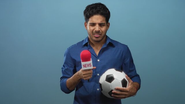 Man holding red microphone and soccer ball in studio while looking down and gripping ball with both hands; nervous interview practice.