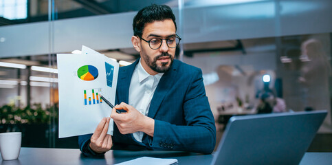 Business professional sits at a table in an office, holding charts and graphs. He discusses data while looking at a laptop screen. The office is bright and modern with plants.
