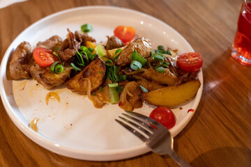 Plate of roasted potatoes with cherry tomatoes and green onions on a wooden table, accompanied by a...
