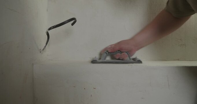 Close-up of a construction worker's hand sanding a plastered surface with a sanding block, creating dust, and then checking the smoothness of the wall with their palm and fingers.