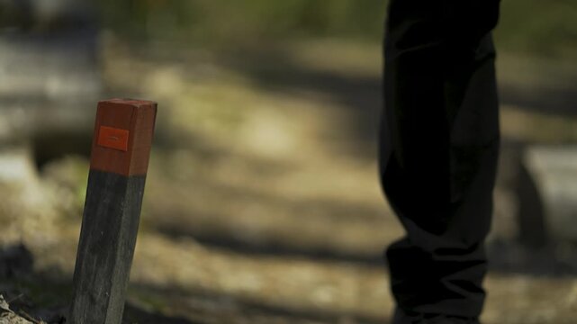 Close-up of a wooden stake with a red trail marker in the foreground and a blurred hiker approaching on a nature path