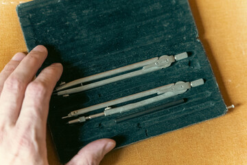 Hand of a person reaching for drafting tools including compasses and pencils inside a black case on a textured orange surface for artistic or technical use