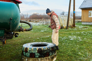 Sanitation worker operating sewage vacuum truck hose near rural cesspit emptier opening during routine waste removal process at homestead.