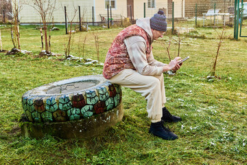 Resting sanitation worker sits on septic tank concrete ring checking smartphone during pause after cesspit maintenance activity.