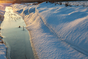 Frozen stream between snow banks during winter thaw, illuminated by low sunlight. Calm winter landscape showing seasonal transition, melting snow and ice with natural copy space