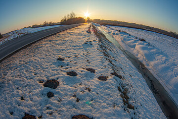 Snowy rural road and frozen ditch at sunrise. Winter landscape with melting snow, strong leading lines and warm sunlight. Seasonal transition in countryside with open copy space