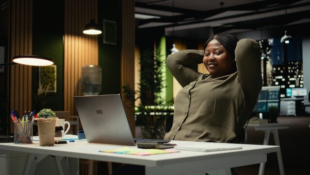 African american woman reclining in a chair in a relaxed pose at midnight, daydreaming during work break. Fulfilled and self assured, reflecting on career success and achievements. Camera A.