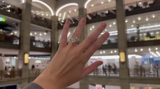 A landscape video shows a hand wearing an engagement ring with a platinum band and oval diamond moving through a spacious shopping gallery with glass ceiling and soft diffused daylight.