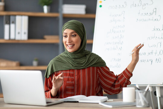 Online Education Concept. Portrait of smiling female arabic teacher in hijab pointing at whiteboard, explaining English grammar rules to students. Excited woman looking at laptop screen, talking