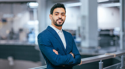 Fototapeta premium A man in a blue suit stands with arms crossed in a bright office. The scene shows an open work area with desks and modern equipment in the background.