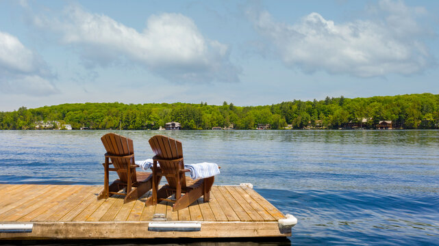 Two wooden Adirondack chairs on a private dock with folded towels overlook a calm Muskoka lake in Ontario, Canada. A pine-lined shore and clear summer sky evoke relaxed cottage life.