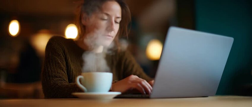 A woman sits in a caf��, absorbed in her laptop, sipping cof