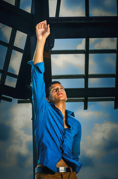 Man Reaching Upward. A man in a blue shirt is looking upwards with one arm extended towards the sky. He stands beneath a metal structure against a backdrop of a partly cloudy sky.