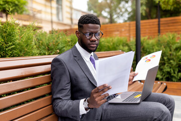 Concentrated african american entrepreneur sitting by office with laptop and reading documents, free space. Confident man in suit checking financial reports, sitting on bench outdoors