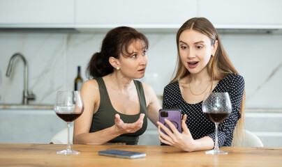 Girls are sitting in kitchen, chatting, reading news on their mobile phone, surfing social...