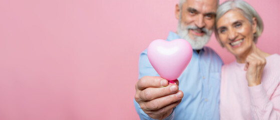 Happy senior couple holding a pink heart shaped balloon pastel background, smiling celebrating love, joyful romantic moment that captures timeless affection, connection relationship goals copy space
