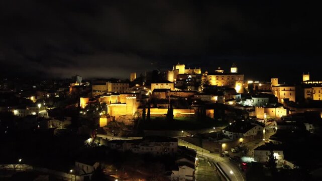 A 4K aerial night video filmed over the illuminated old town of Caceres in Extremadura, Spain. The drone moves slowly sideways, revealing the beautifully lit medieval historic center and stone