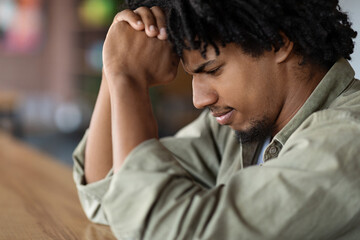Fototapeta premium Sad serious concentrated young african american curly male praying or thinking about solving problem at table in cafe interior, profile, close up, cropped. Dream, facial expression, gesture and hope