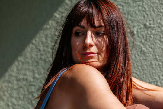 Young woman with long dark hair and natural makeup poses gracefully against a textured green wall, showcasing a relaxed and contemplative mood in a serene environment