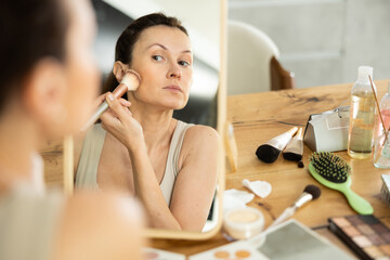 Calm middle-aged woman applying blush on her cheeks by means of makeup brush sitting in front of the mirror