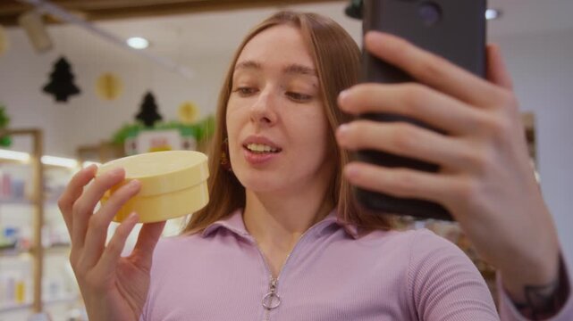 Rack focus from smartphone in hand of smiling female influencer reviewing skincare product holding cosmetic jar while recording sponsored content at beauty store