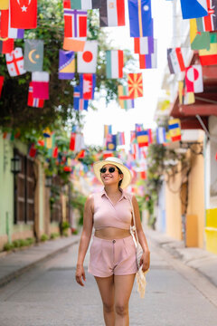 Young happy smiling Latin woman strolling joyfully under vibrant international flags in Getseman&iacute;, Cartagena, Colombia. She wears a pale pink summer outfit, sun hat and sunglasses.